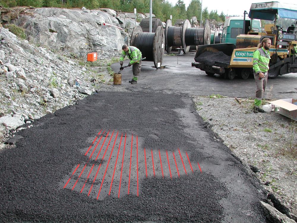 Santier cu asfalt proaspat asternut peste cablul de degivrare, cu traseul cablului marcat cu rosu si cilindrul compactor in asteptare